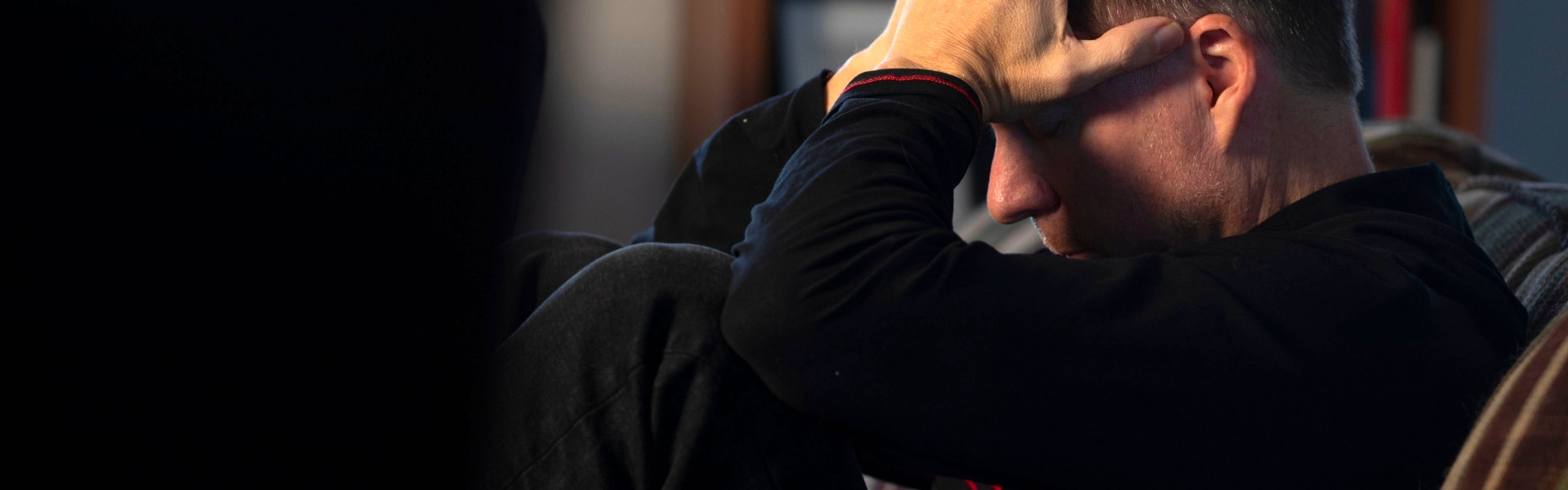 Man zit ineengedoken op de bank met zijn hand tegen het voorhoofd, beeld bij depressie en somberheid.