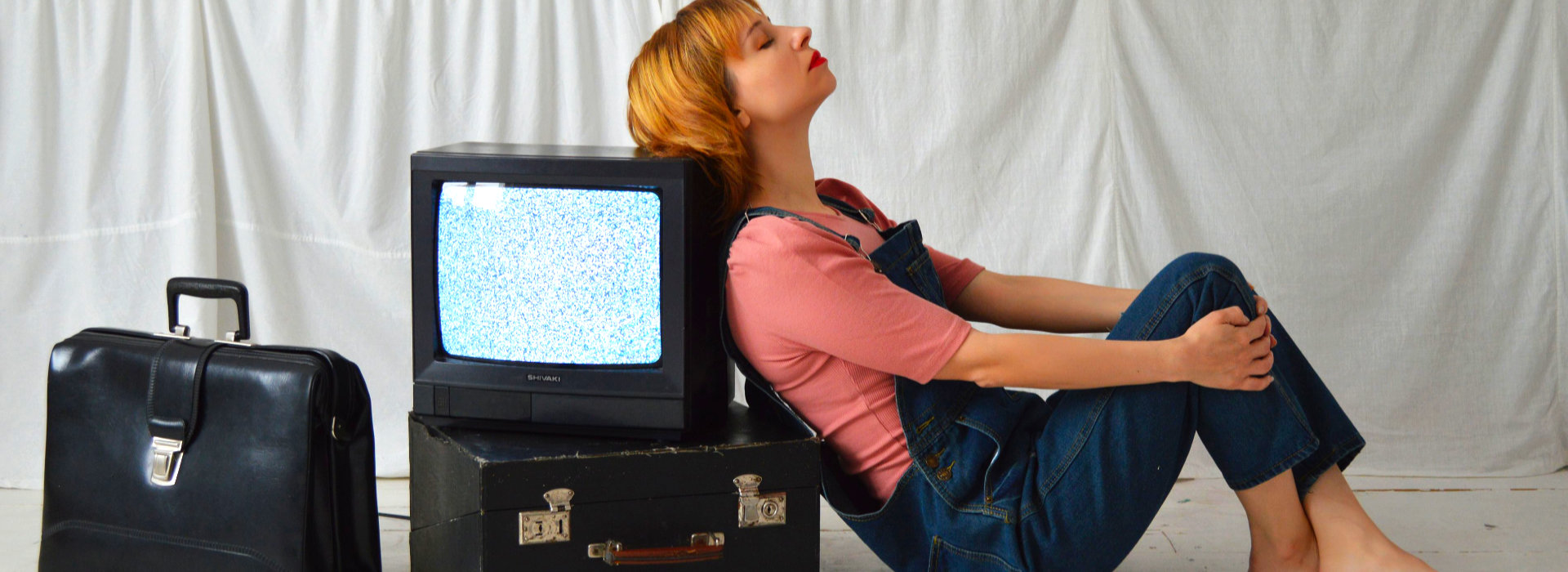 Vrouw leunt vermoeid tegen een oude televisie en koffers, beeld bij coaching en therapie rond verleden loslaten bij Spreek met Freek.