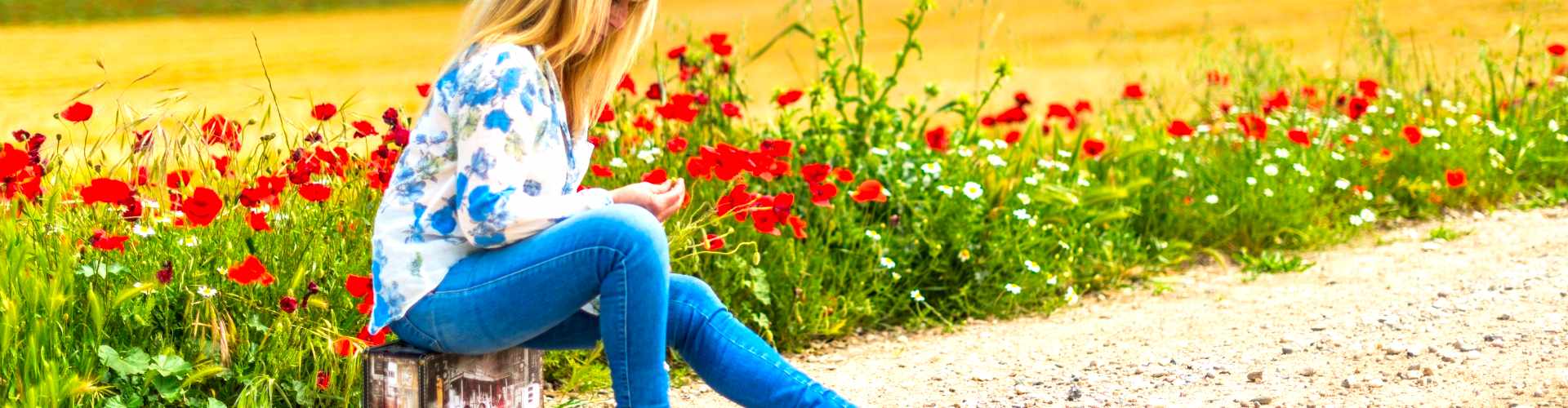Vrouw zit op een koffer langs een weg tussen bloemen, beeld bij coaching en therapie rond verleden loslaten en emotionele bagage bij Spreek met Freek.